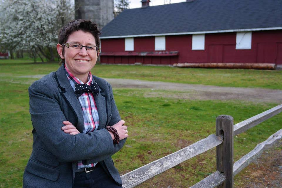 A woman with short brown hair and pale skin leans against a fence and smiles next to a barn.