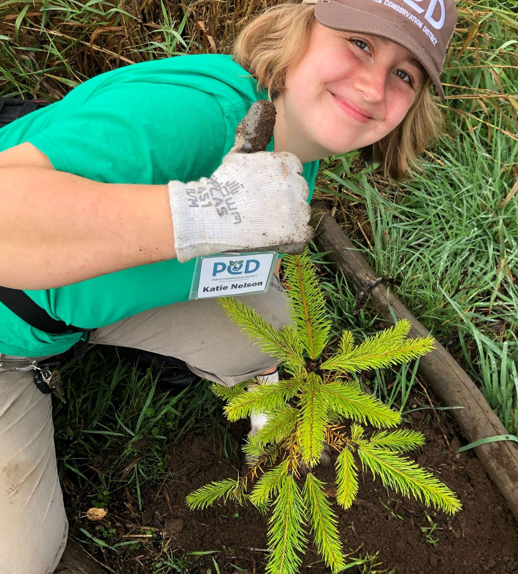A long blonde haired woman poses with a thumbs up next to a freshly planted tree