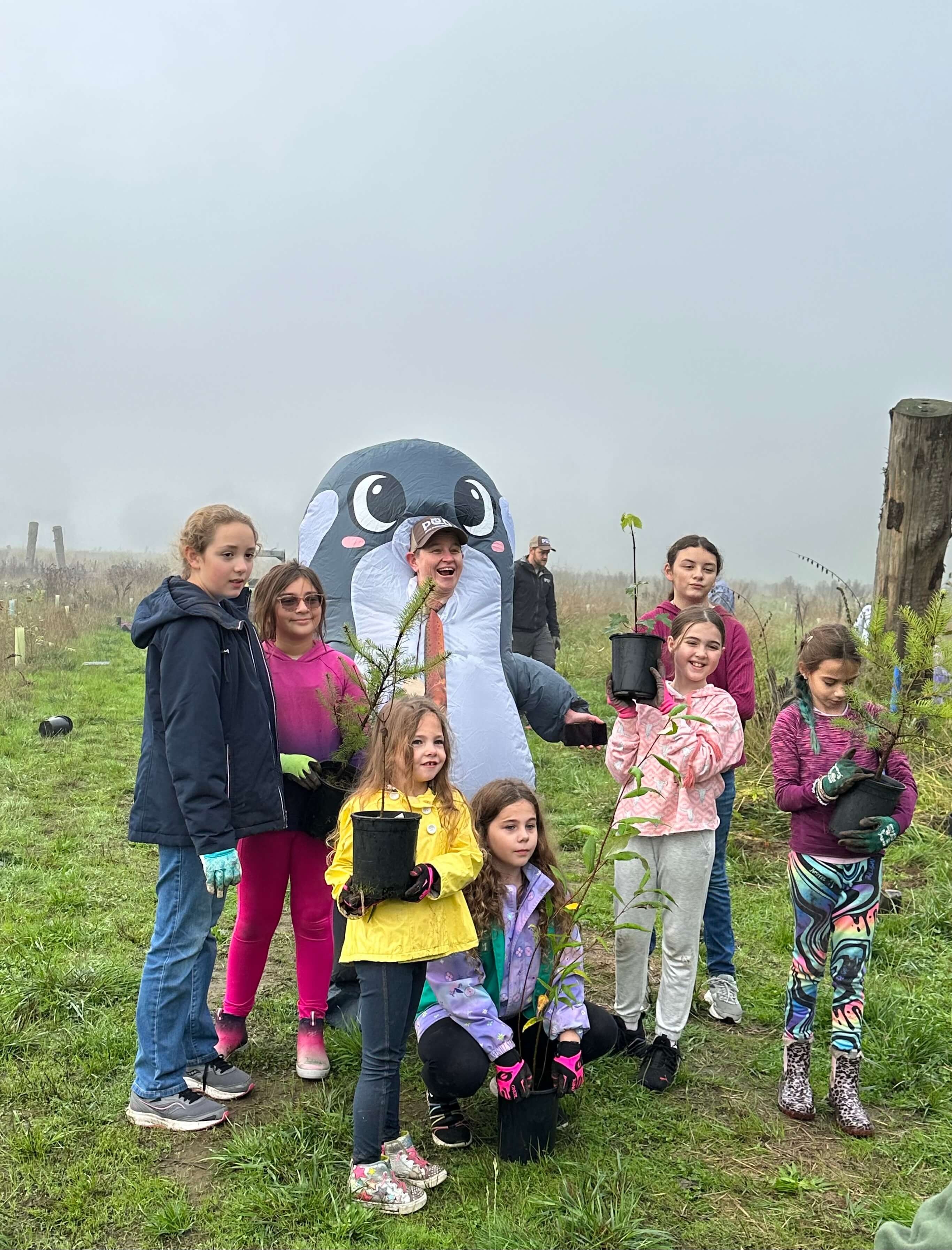 Seven children holding trees posing with Executive Director Dana Coggon in an Orca costume.