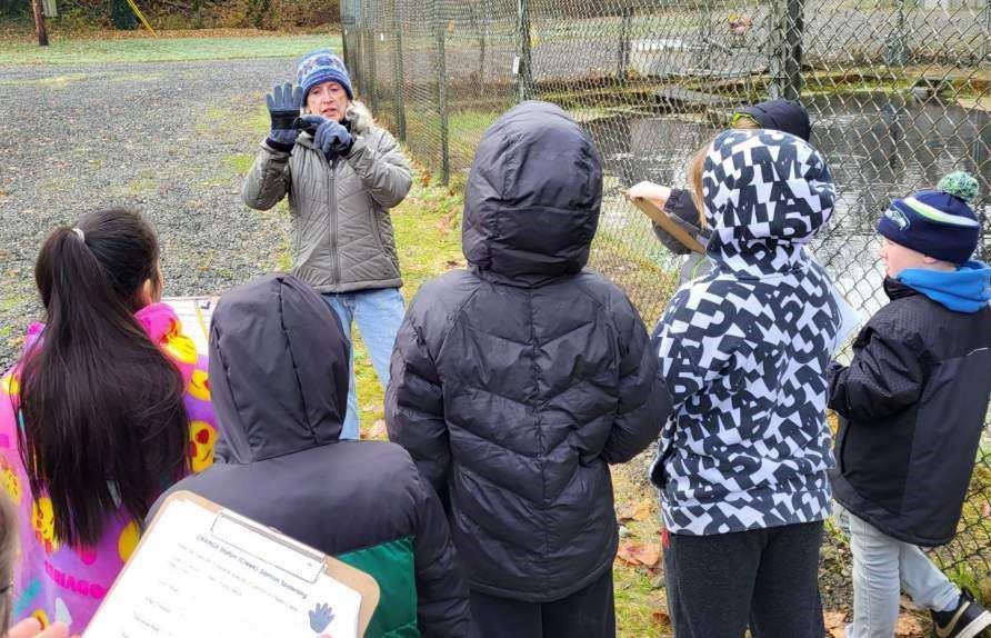 An education docent holding up their hand and pointing to their palm, while students in coats listen