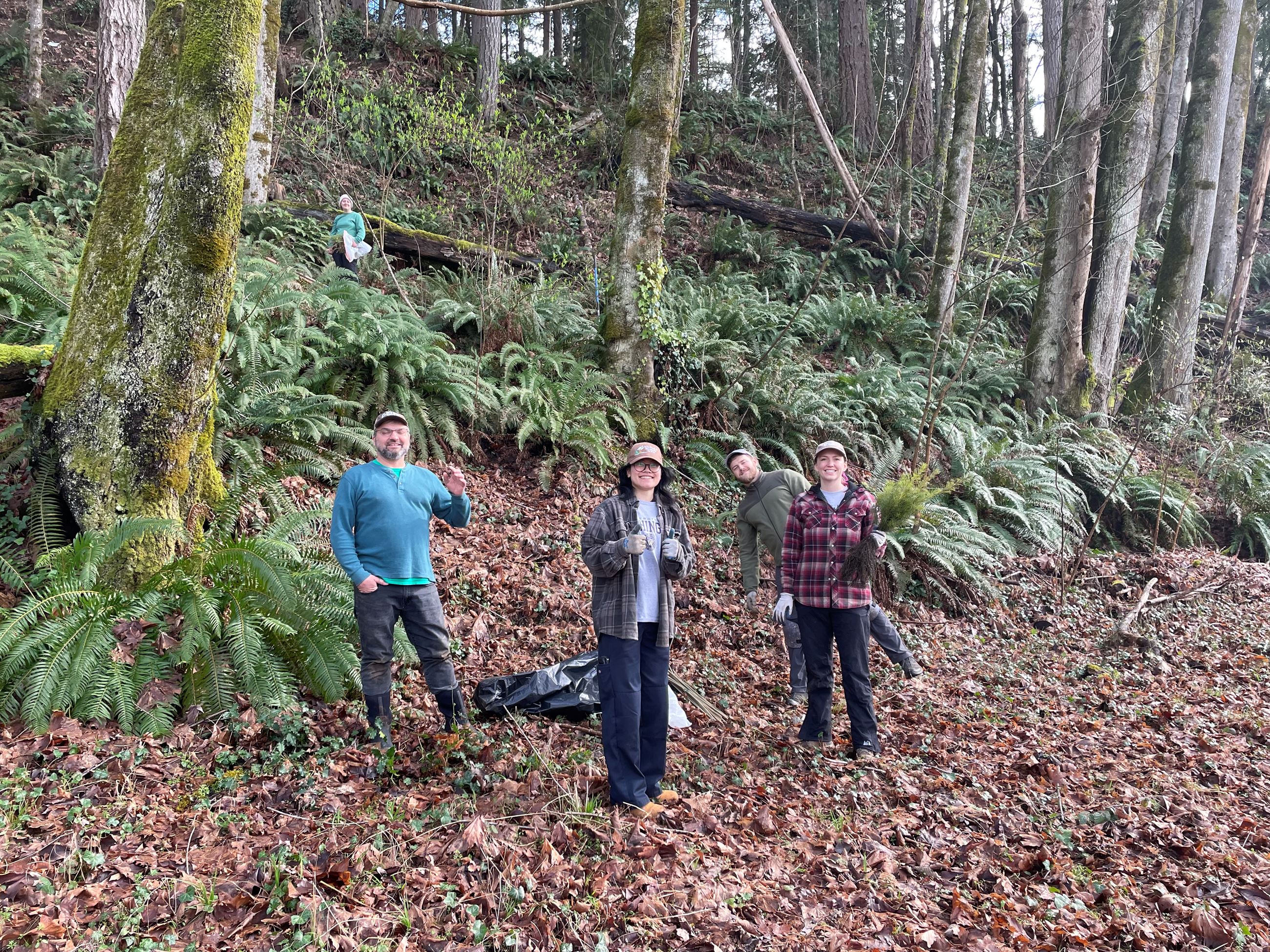 Three staff members stand in a forested area with trees behind them and leaves covering the ground.