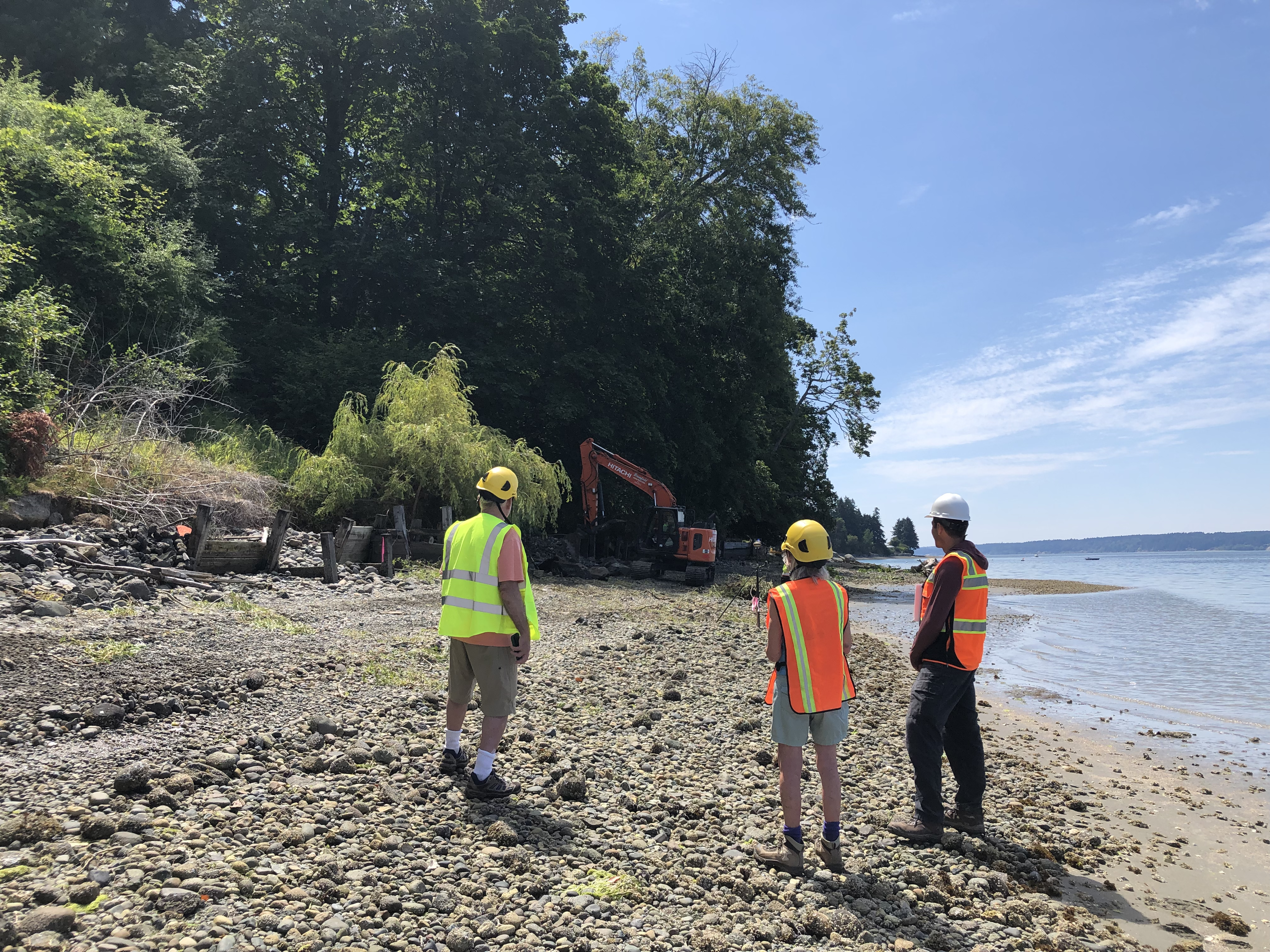 Three people in safety vests stand on the beach watching the excavators in the background working on