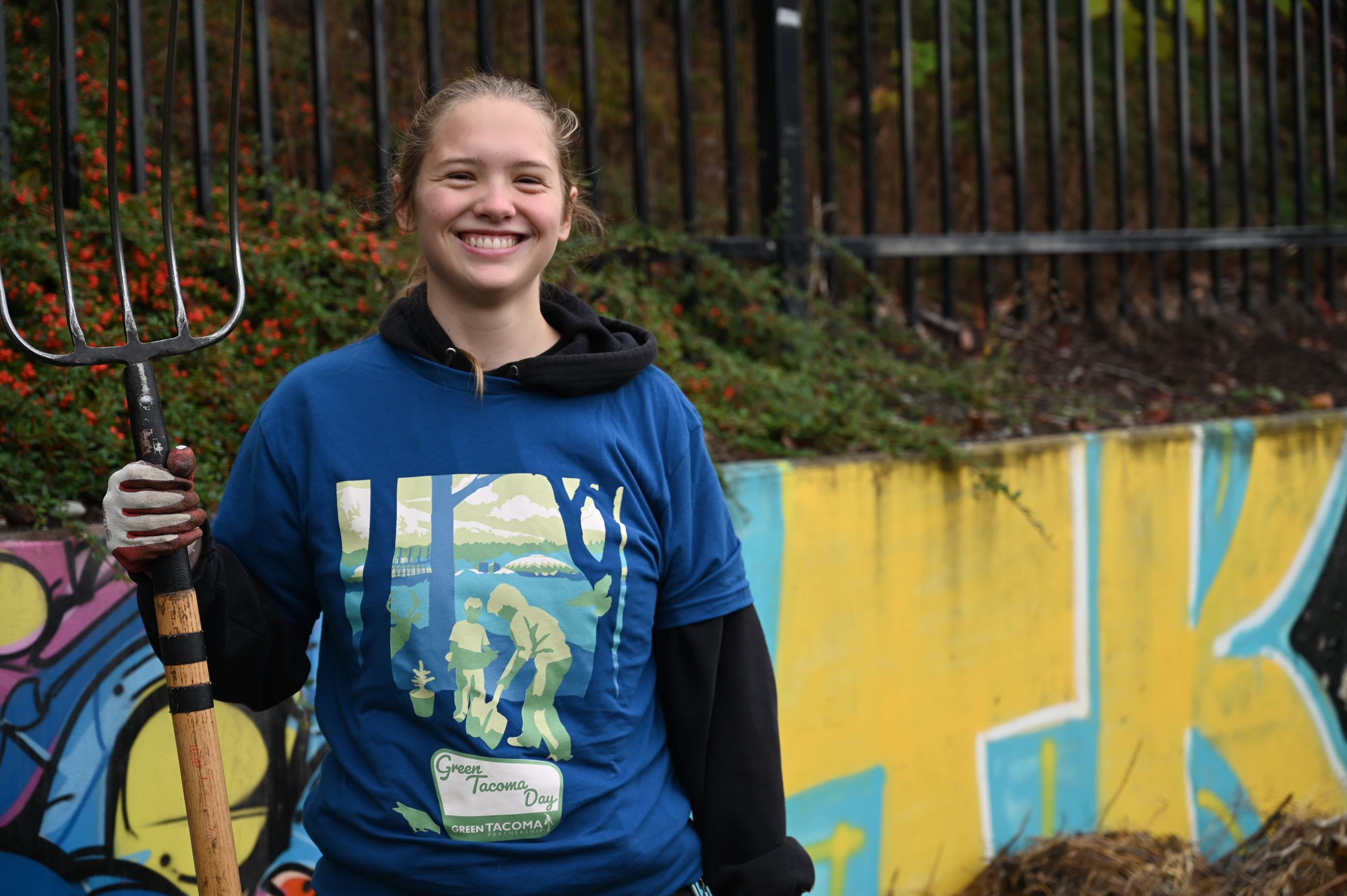 a volunteer wearing a green Tacoma day shirt posing with a pitchfork and smiling at the camera